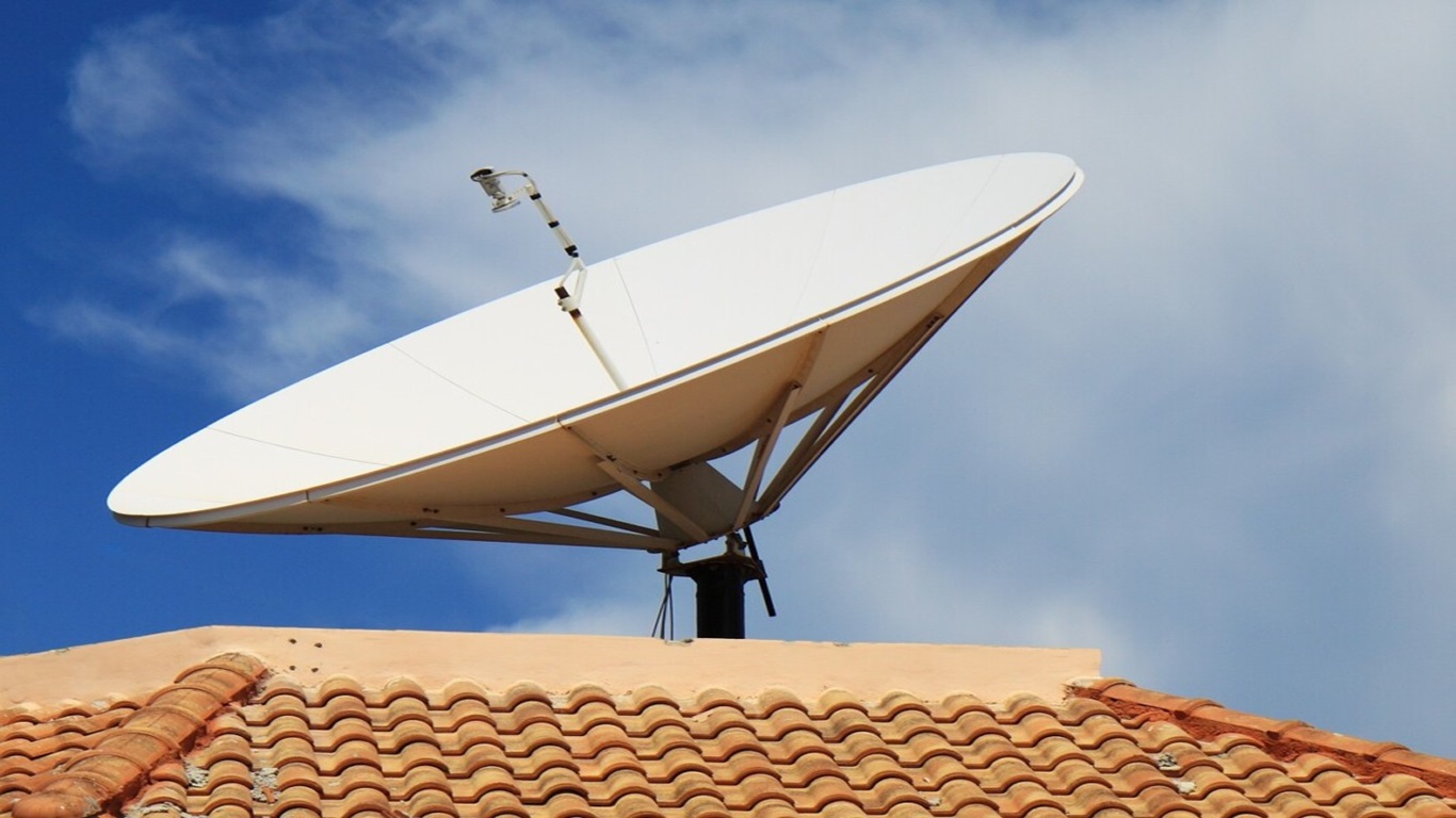 Antena parabólica branca fixada sobre um telhado de cerâmica, direcionada para o céu azul.