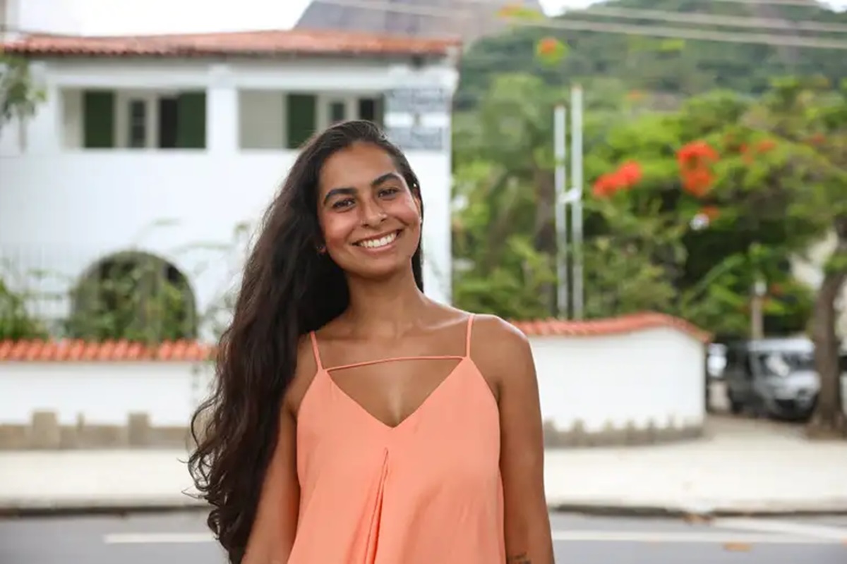 Jovem sorridente de pele morena e longos cabelos escuros em frente a uma casa branca e vegetação exuberante.