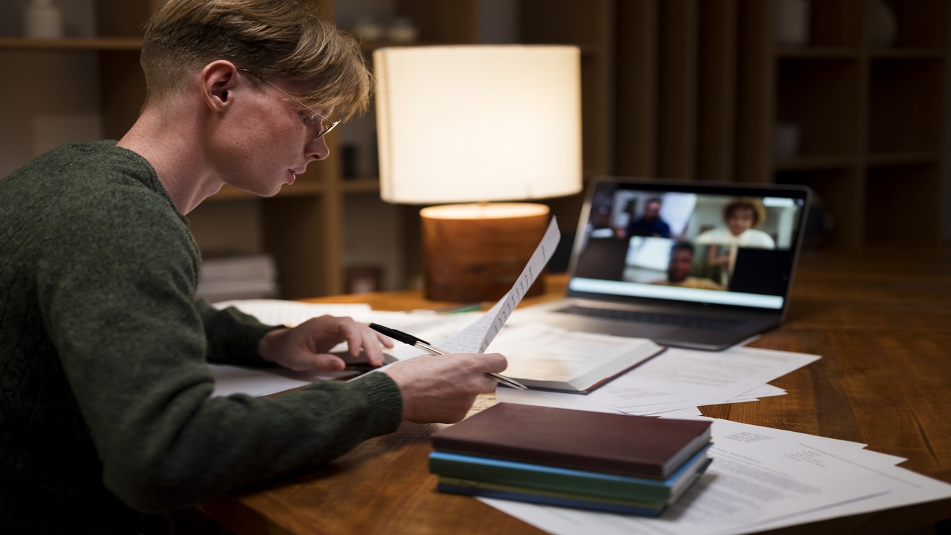 Jovem estudando em sala virtual com documentos e computador à noite