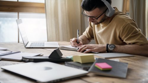 Estudante de óculos e fones de ouvido anota em caderno enquanto assiste aula online em laptop sobre mesa de madeira com outros materiais de estudo