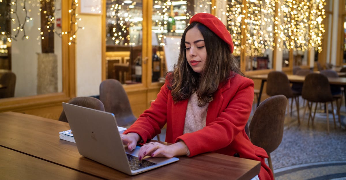 Mulher com cabelo longo, usando um casaco vermelho, digitando em laptop em um café com luzes decorativas.