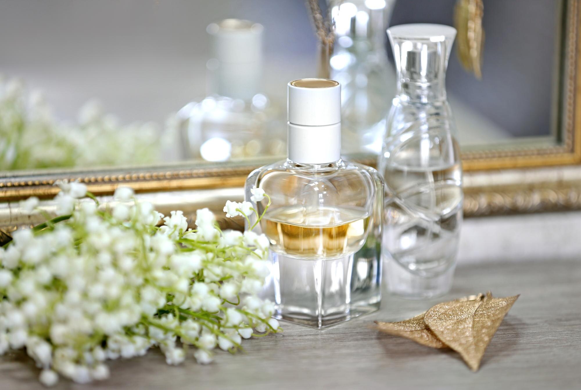 Frasco de perfume dourado sobre penteadeira de madeira, ao lado de flores brancas de lírio-do-vale e espelho com moldura dourada
