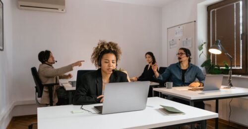 Grupo de pessoas trabalhando em escritório, com fones de ouvido e computadores.