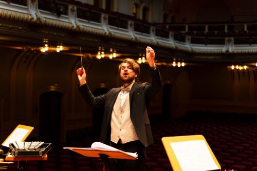 Maestro de fraque ergue a batuta ao reger uma orquestra em uma sala de concerto ornamentada e iluminada