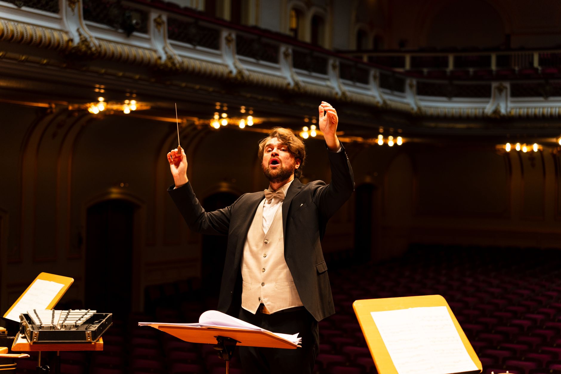 Maestro de fraque ergue a batuta ao reger uma orquestra em uma sala de concerto ornamentada e iluminada