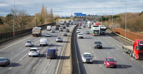 Imagem de uma rodovia com vários carros e caminhões em circulação sob céu nublado.