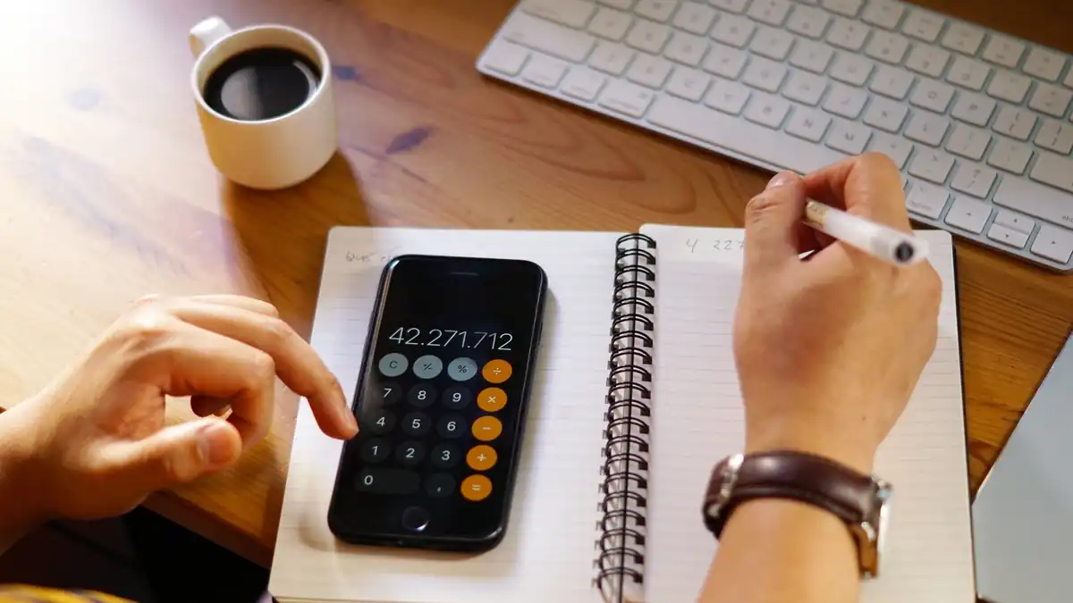Maos fazendo calculos no celular e anotacoes em caderno.