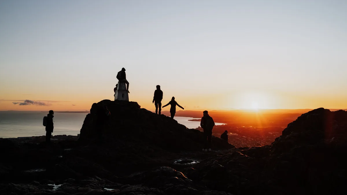 Silhuetas de pessoas no alto de um morro rochoso ao pôr do sol, com o mar e a cidade ao fundo