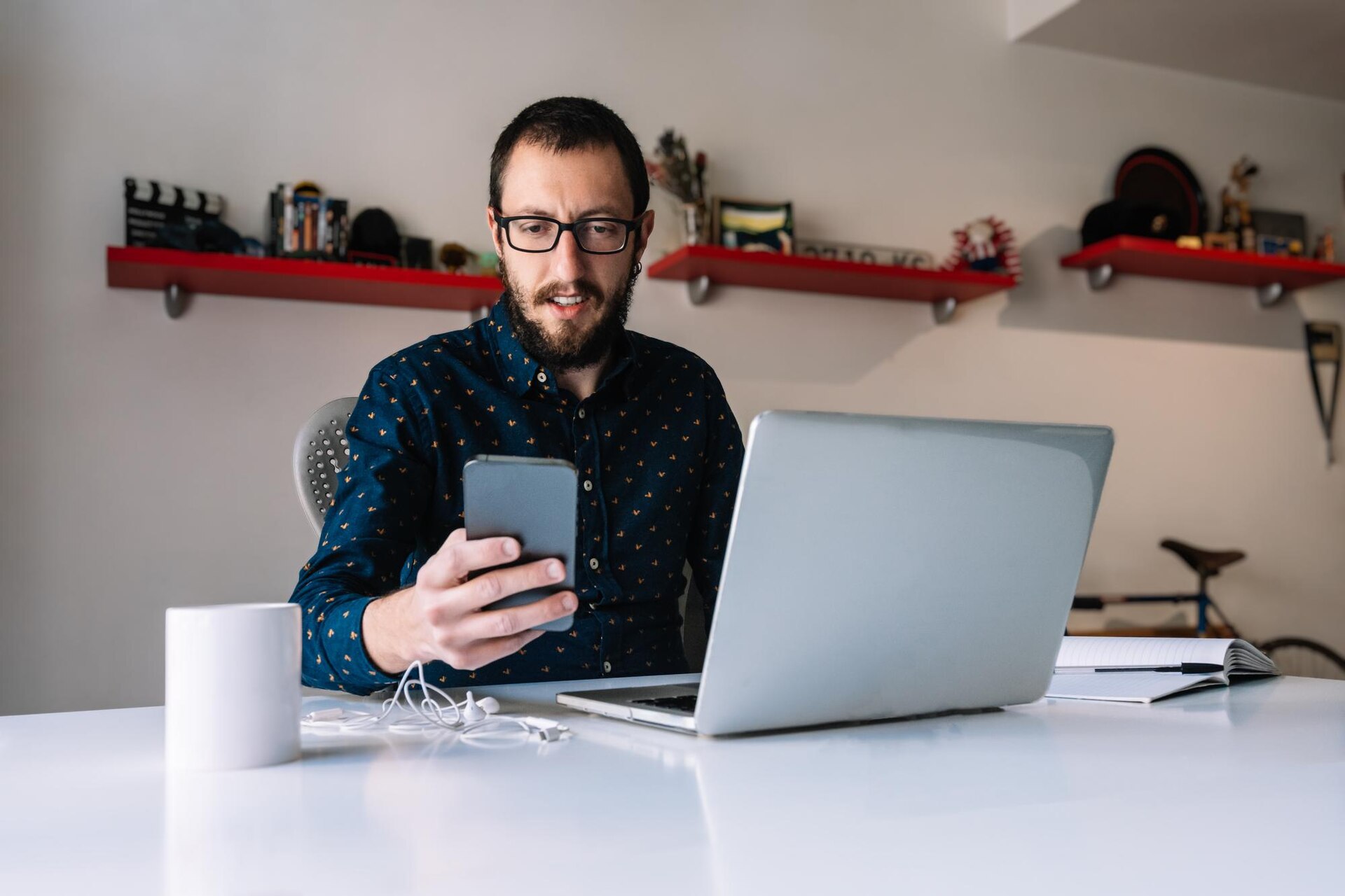 Homem de óculos e barba segura um celular enquanto trabalha diante de um notebook aberto sobre uma mesa branca